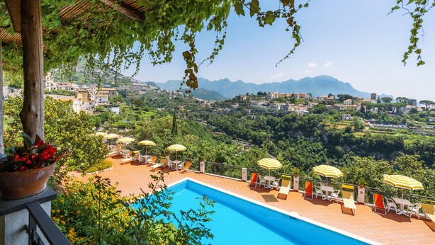 Una piscina con sillones de playa y sombrillas, rodeada de vegetación y vistas a un pueblo y montañas.