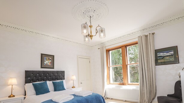 A bedroom with a large bed, two bedside tables, a chandelier, a window with curtains, and a chair. - Mary Ann Villa (Inverness)