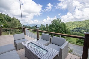 A deck with outdoor furniture and a glass-top table, overlooking a road and green hills.