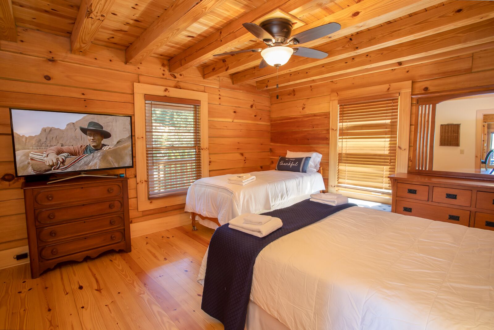 A log cabin bedroom with two beds, a ceiling fan, wooden floors, and a television on a dresser.
