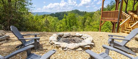 A fire pit surrounded by Adirondack chairs, with a wooden deck and a scenic mountain view in the background.