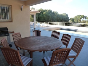 Mesa redonda de madeira com seis cadeiras, churrasqueira e vista para a piscina.