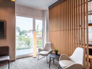 A modern hotel room with a large window, a seating area with two chairs and a small table, and a wooden slat partition. - B&B Hotel Augsburg-Nord (Augsburg)