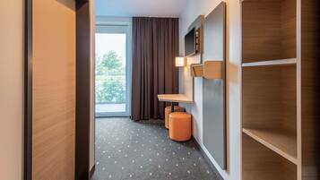 Un pasillo de hotel con suelo de madera, un ventanal con cortinas, una pequeña mesa con lámpara y estanterías empotradas.