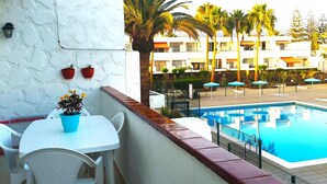 Un balcon avec une table et des chaises blanches, une plante en pot, et un espace piscine agrémenté de parasols et de palmiers en arrière-plan.