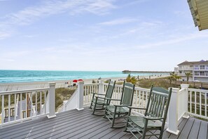 A wooden deck with rocking chairs overlooking a beach and pier.