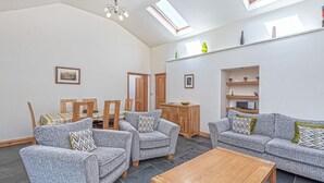 A spacious living room with grey sofas, wooden chairs, a wooden coffee table, and a red rug. There are skylights, a wooden cabinet, and shelves with decorative items.