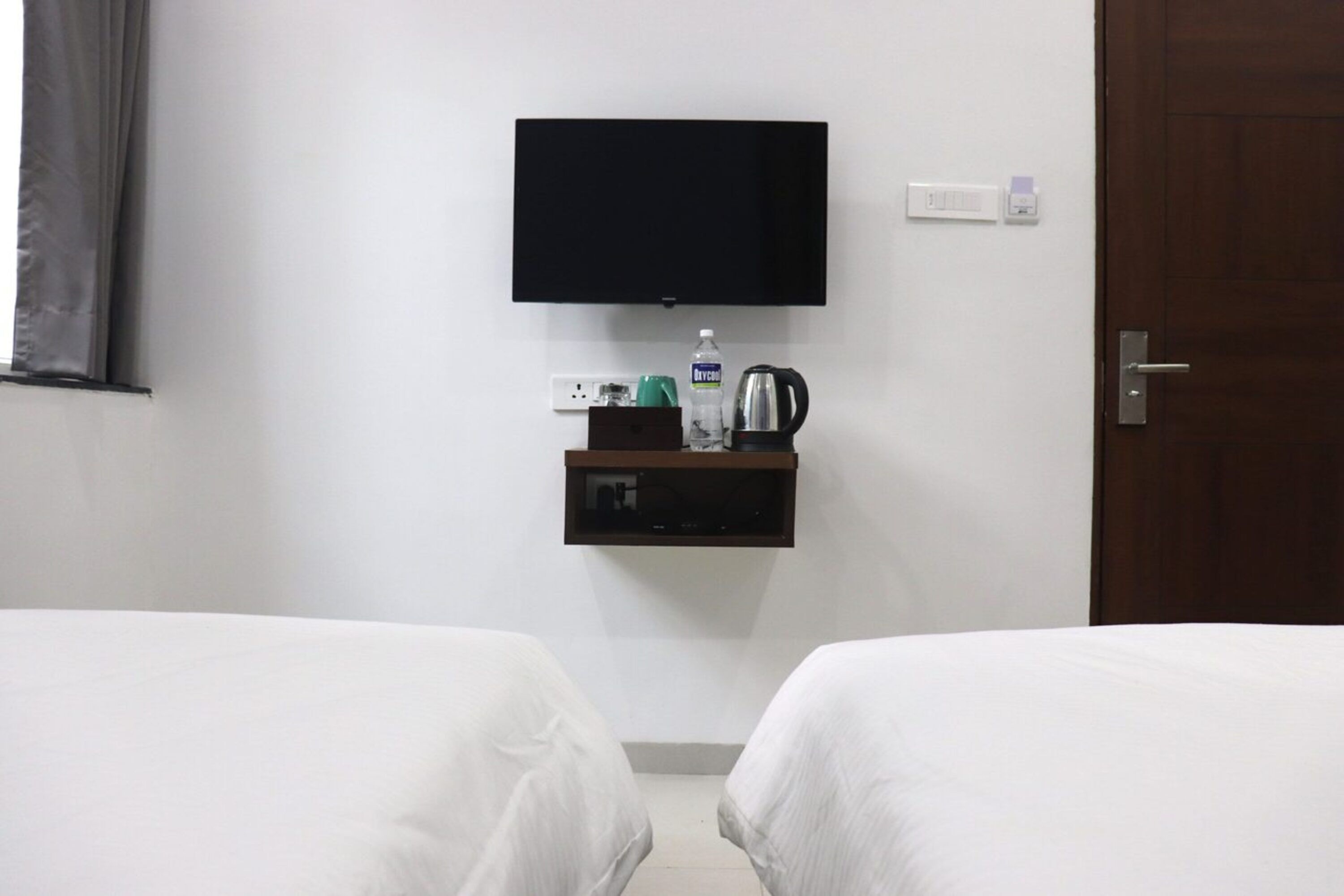 Ein Hotelzimmer mit zwei Betten, einem an der Wand befestigten Fernseher, einer kleinen Ablage mit Wasserkocher, einer Wasserflasche und einer Steckdose.