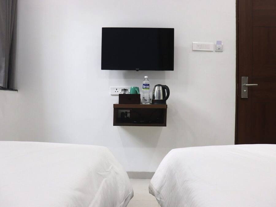 Habitación de hotel con dos camas, un televisor montado en la pared, un estante pequeño con una tetera, una botella de agua y un enchufe.