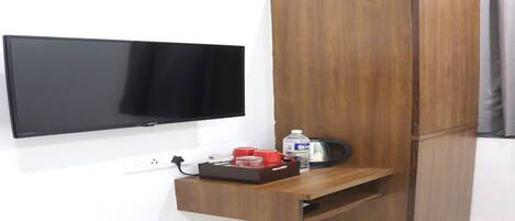 A room with a flat-screen TV mounted on the wall, a wooden wardrobe with a built-in shelf, and a small table with a bottle and glasses.