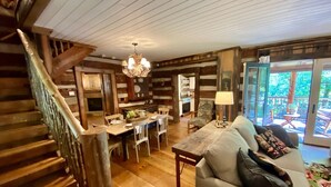 A rustic cabin interior with a wooden staircase, a dining area, and a sofa with a view to an outdoor deck.
