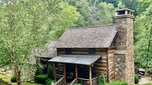 A rustic log cabin with a stone chimney, a porch with a metal roof, and a stone pathway leading to the entrance.