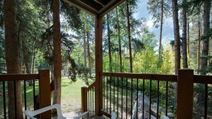Eine Holzterrasse mit zwei weißen Plastikstühlen, umgeben von hohen Bäumen und einer Rasenfläche.