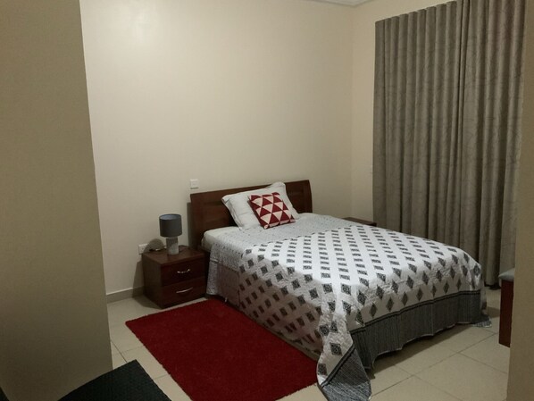 A bedroom with a bed, a nightstand, a lamp, a red rug, and a window with curtains. - Charming 3-bed Apartment Airport Residential Accra (Accra)