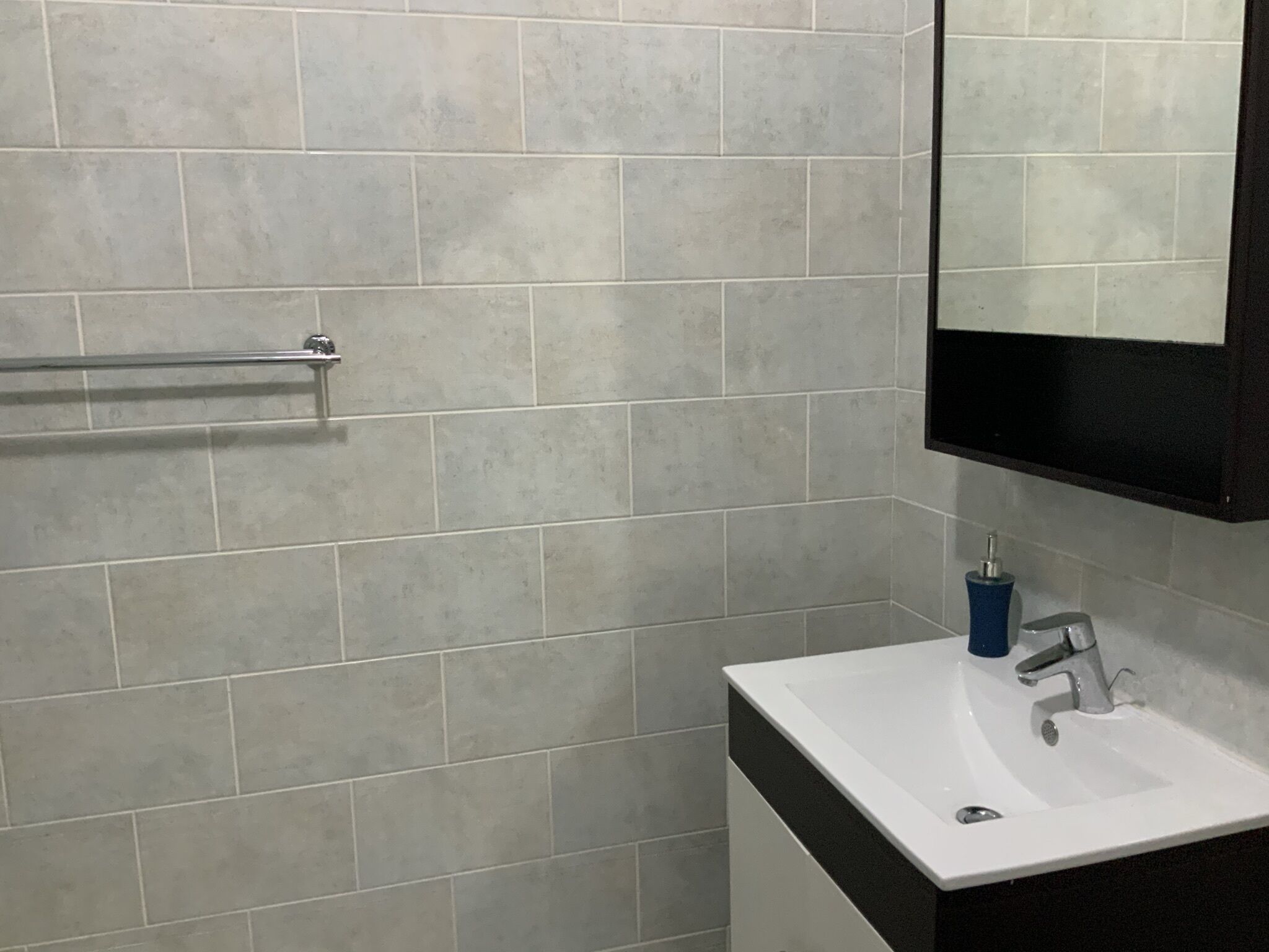 A bathroom with grey tiled walls, a white sink, a blue soap dispenser, and a metal towel rack.