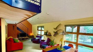 A spacious living area with a high ceiling, a staircase, a dining table with chairs, and a large window with a view of greenery. - Hummingbird Hideaway (Galeton)