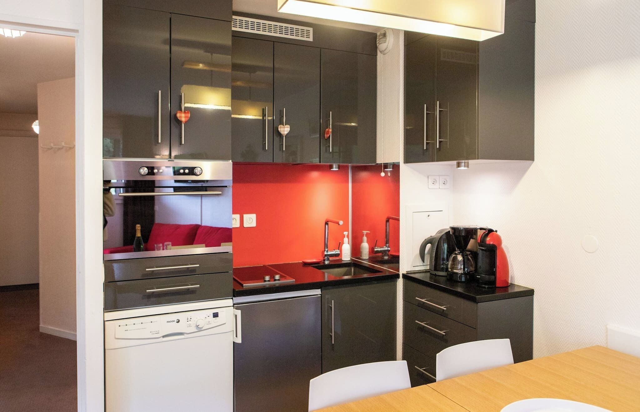 A compact kitchen with modern appliances, a red accent wall, and a dining area with a wooden table and white chairs.