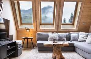 Un dormitorio tipo loft acogedor con un sofá gris grande, una mesa de centro de madera y un escritorio con una lámpara. Tres tragaluces ofrecen vistas a un paisaje montañoso.