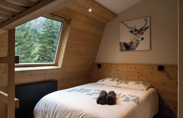 Un dormitorio con techo de madera, una cama con cubrecama blanco y dos toallas enrolladas, una ventana con vista a árboles, y una obra de arte de un ciervo en la pared.