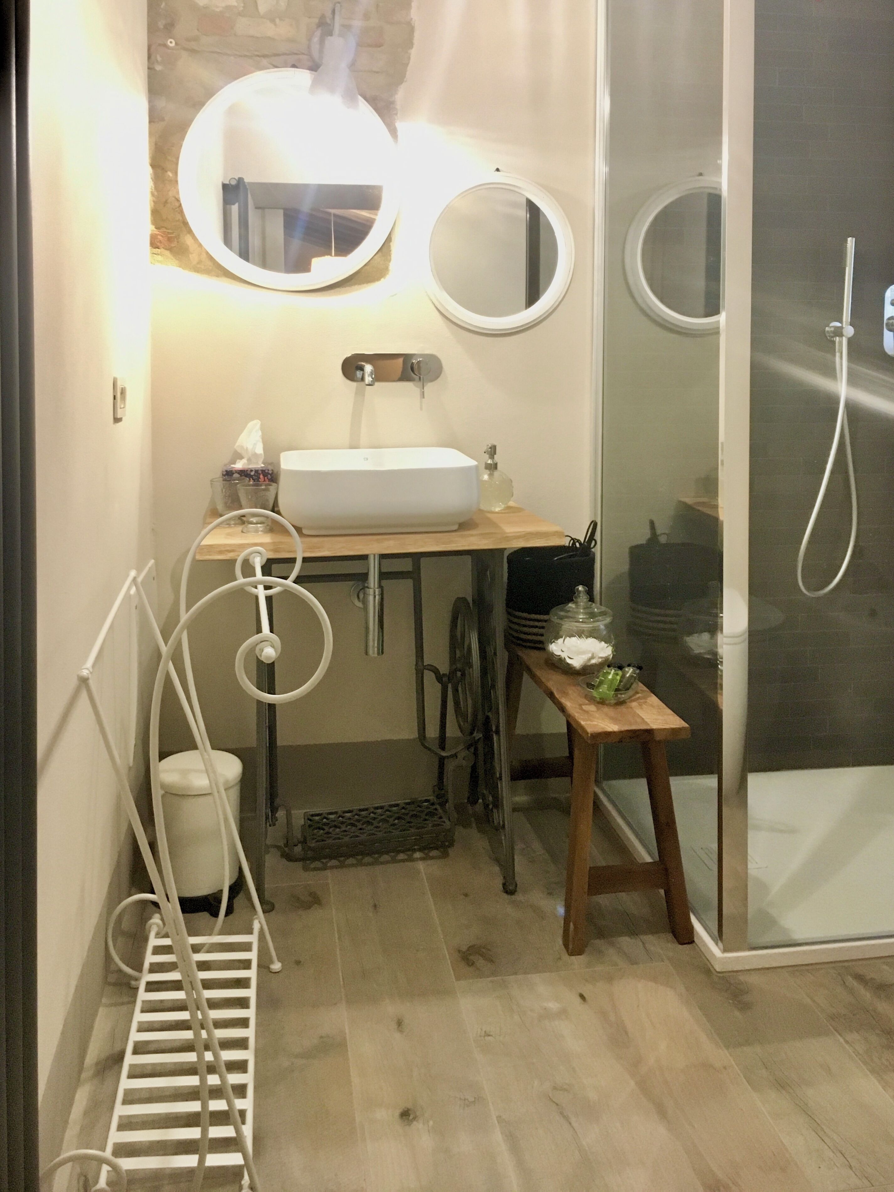 Un baño pequeño con un lavamanos blanco, dos espejos montados en la pared, una ducha y una pequeña mesa de madera con artículos de tocador.