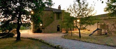 Un edificio rústico de piedra con techo inclinado, un camino de grava que conduce a él, y un parque infantil con un tobogán.