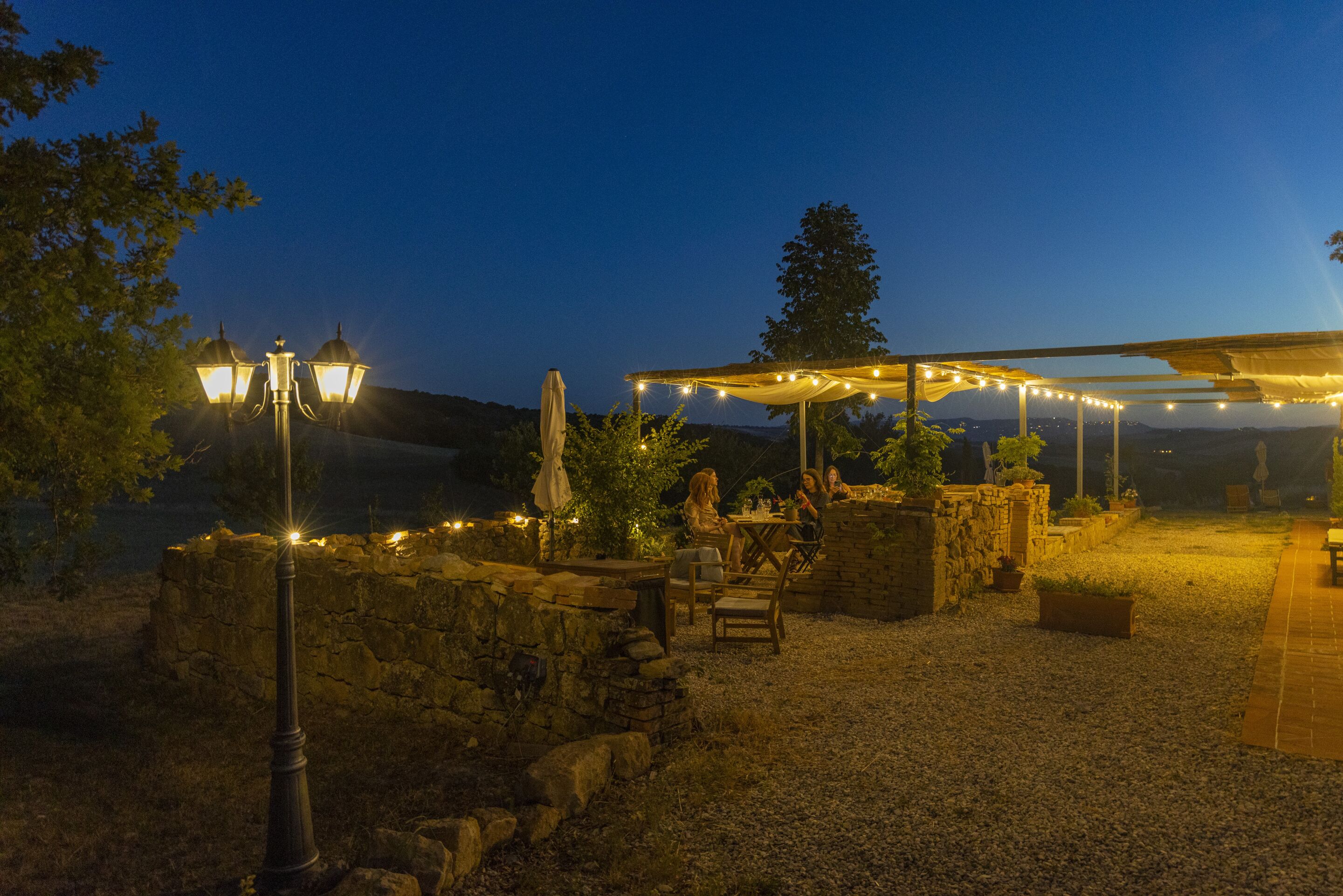 Un espace extérieur aménagé en terrasse, avec des murs en pierre, une pergola illuminée de guirlandes lumineuses et une table entourée de chaises.