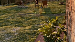 Um sino com pássaros em uma estrutura metálica, um pôr do sol ao fundo e um gramado com cadeiras.