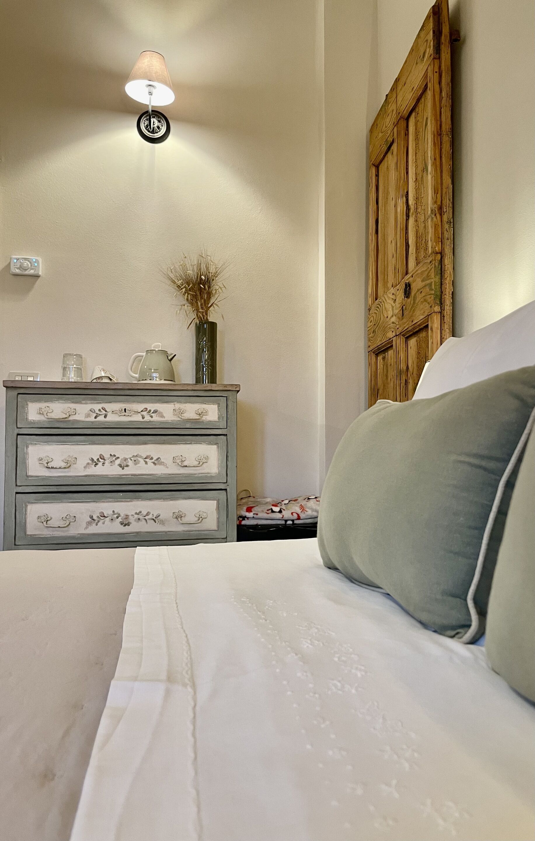Een slaapkamer met een bed, een dressoir met lades, een lamp, een houten deur en een vaas met gedroogde planten.