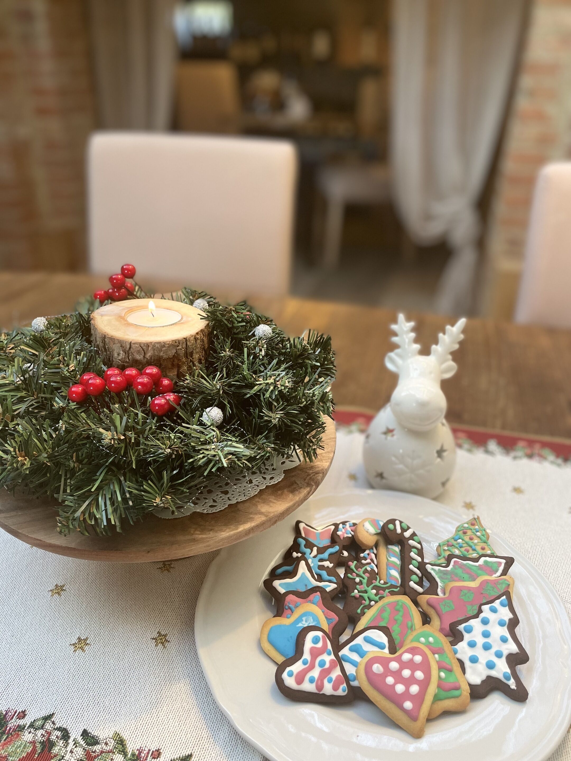 Une table ornée d’un centre de table en bois, composé d’une bougie et de quelques éléments de verdure, ainsi que d’une assiette remplie de biscuits décorés.