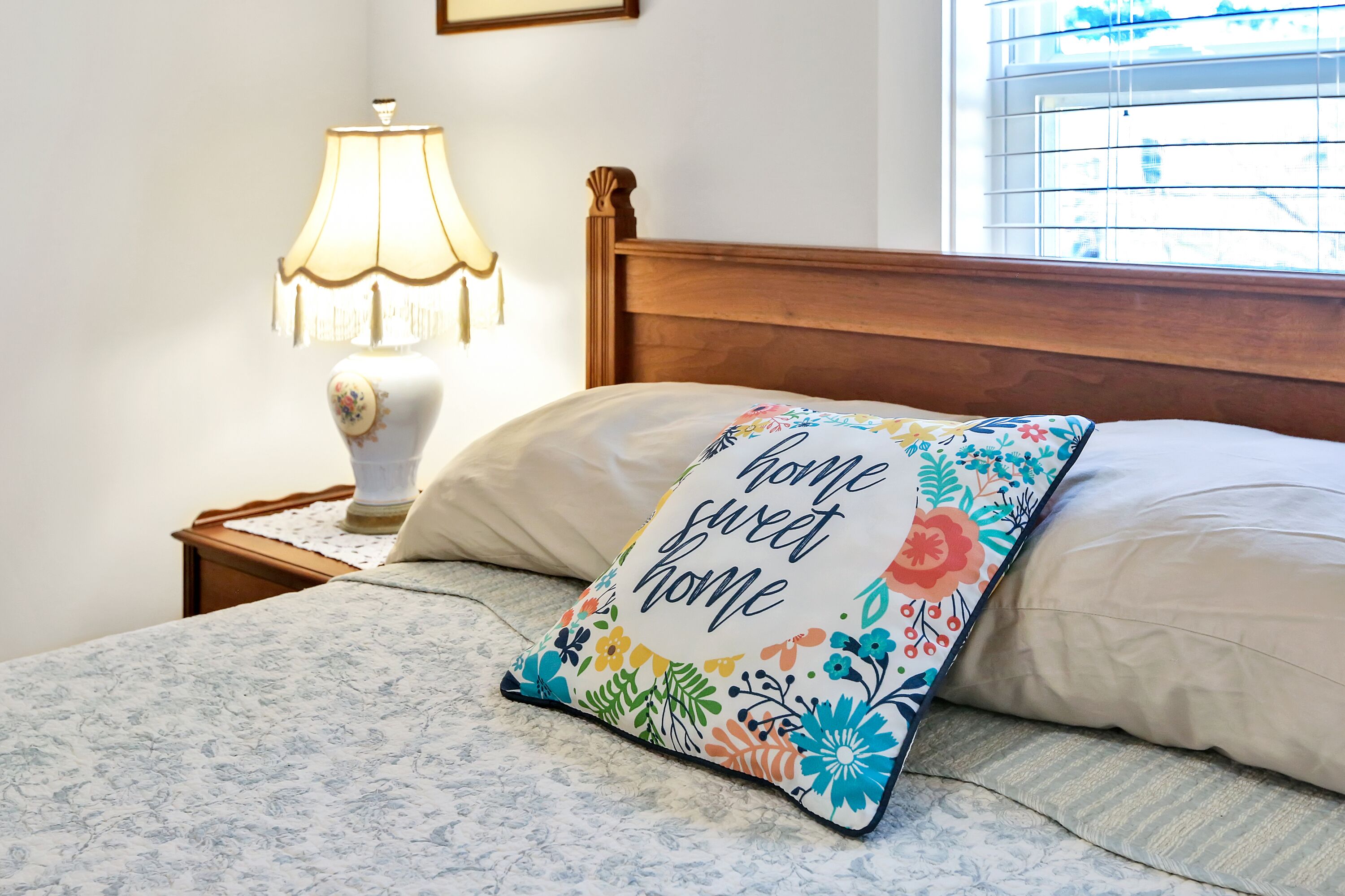 Una cama bien hecha con un cojín floral, una lámpara de noche y una ventana con persianas.