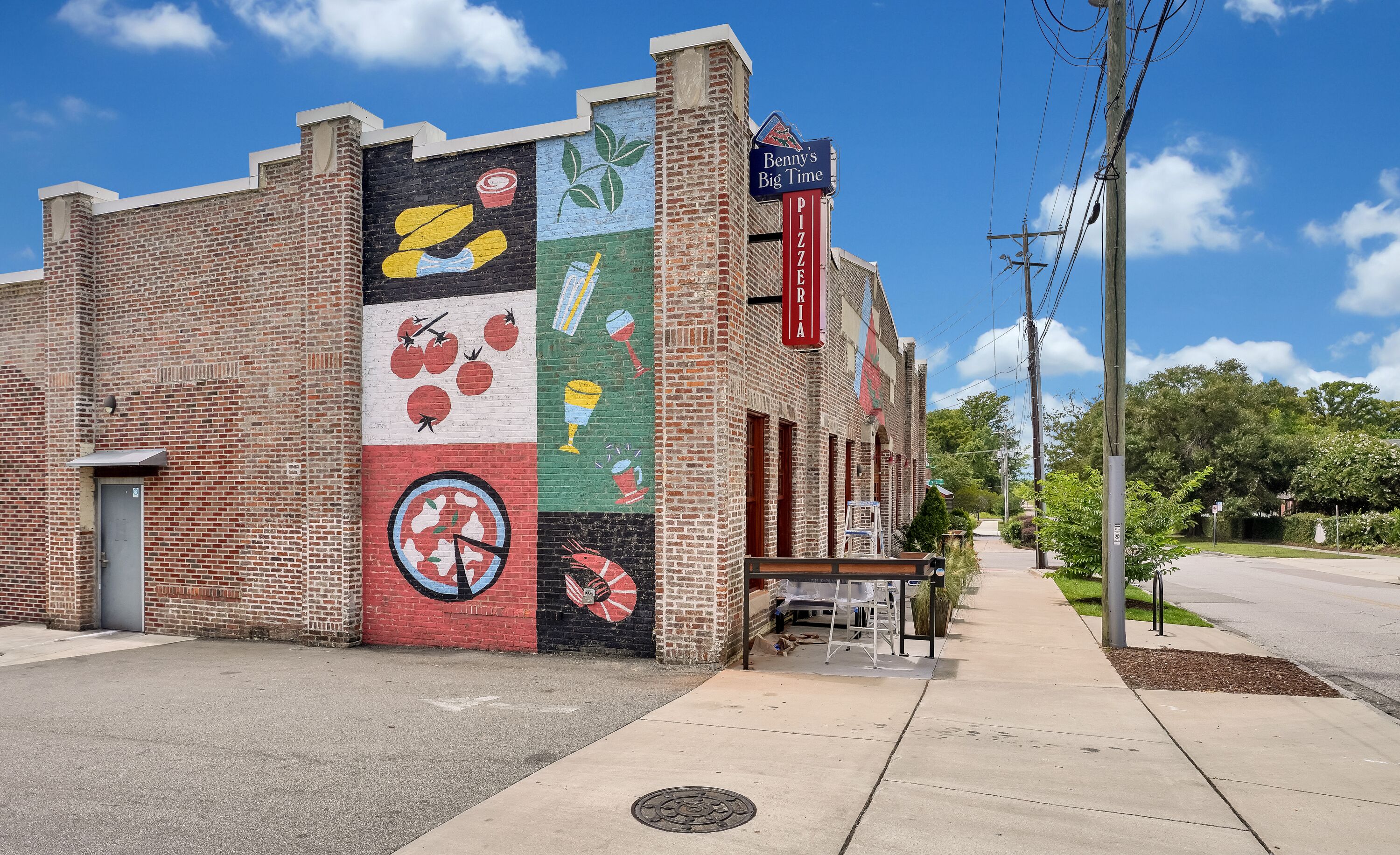 Un bâtiment en briques orné d’une fresque sur le côté, une enseigne indiquant « Benny’s Big Time Pizza », et un trottoir devant.