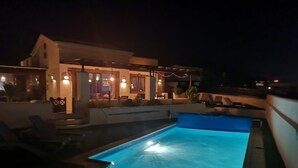 A swimming pool with a blue interior, surrounded by lounge chairs and a building with outdoor seating. - Luxurious 2 Bed Villa (Montaña Roja, Playa Blanca)