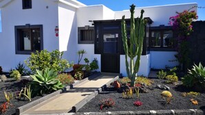 A white building with a black door and windows, surrounded by a garden with various plants and a cactus. - Luxurious 2 Bed Villa (Montaña Roja, Playa Blanca)
