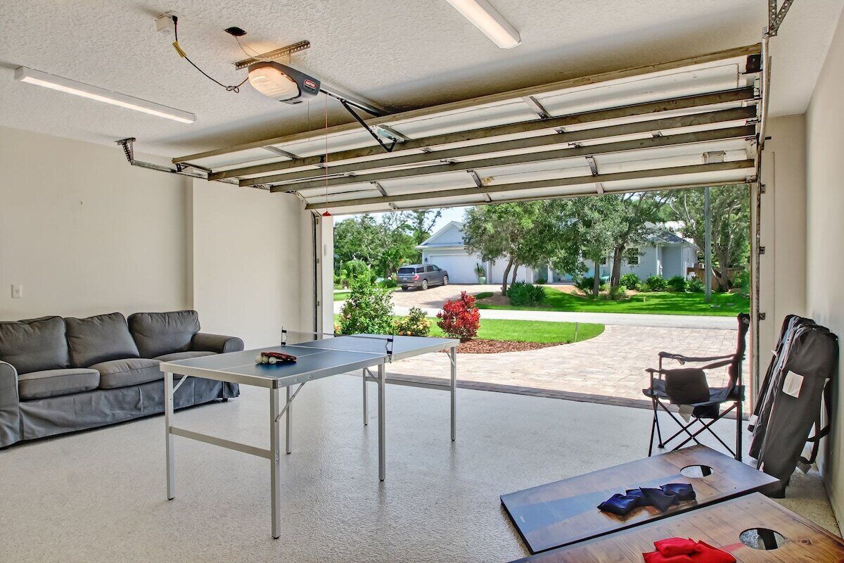A garage with a table and a couch, a folding chair, and a view of a residential area.