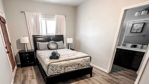 A bedroom with a bed, two bedside tables, a window with curtains, a door, and a small bathroom visible through the door. - Centrally Located River Home with Mountain Views! (Fort Mohave)