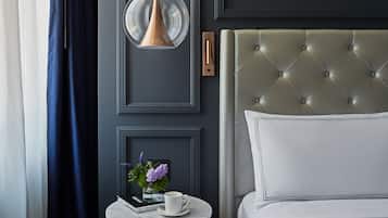 A neatly arranged hotel room with a tufted headboard, a round table with a book and a vase of flowers, and a hanging pendant light.
