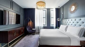Ein modernes Hotelzimmer mit einem großen Bett, einem Fernseher, einer Chandelüster und Blick auf die Stadt durch die Fenster.