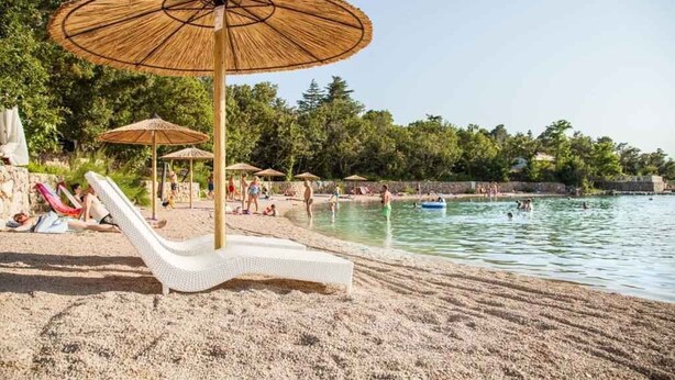 En strand med hvide liggestole og parasoller, et poolområde samt folk der svømmer.