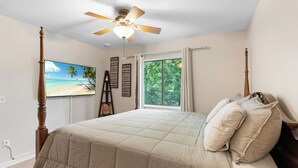 Une chambre avec un lit, un ventilateur de plafond, une fenêtre avec des rideaux, un tableau représentant une plage et une citation inspirante sur le mur.