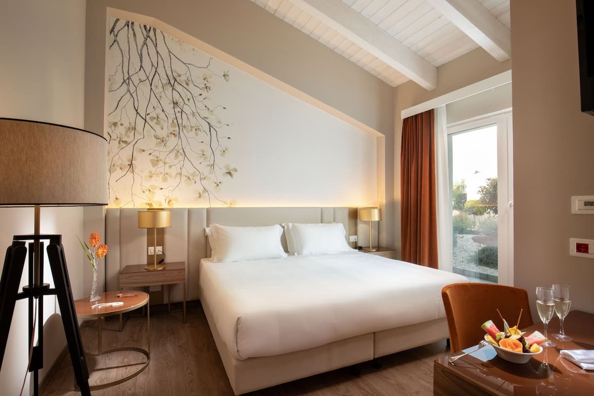 Quarto de hotel com uma cama grande, uma escrivaninha com abajur, um vaso com flores e vista para o exterior.
