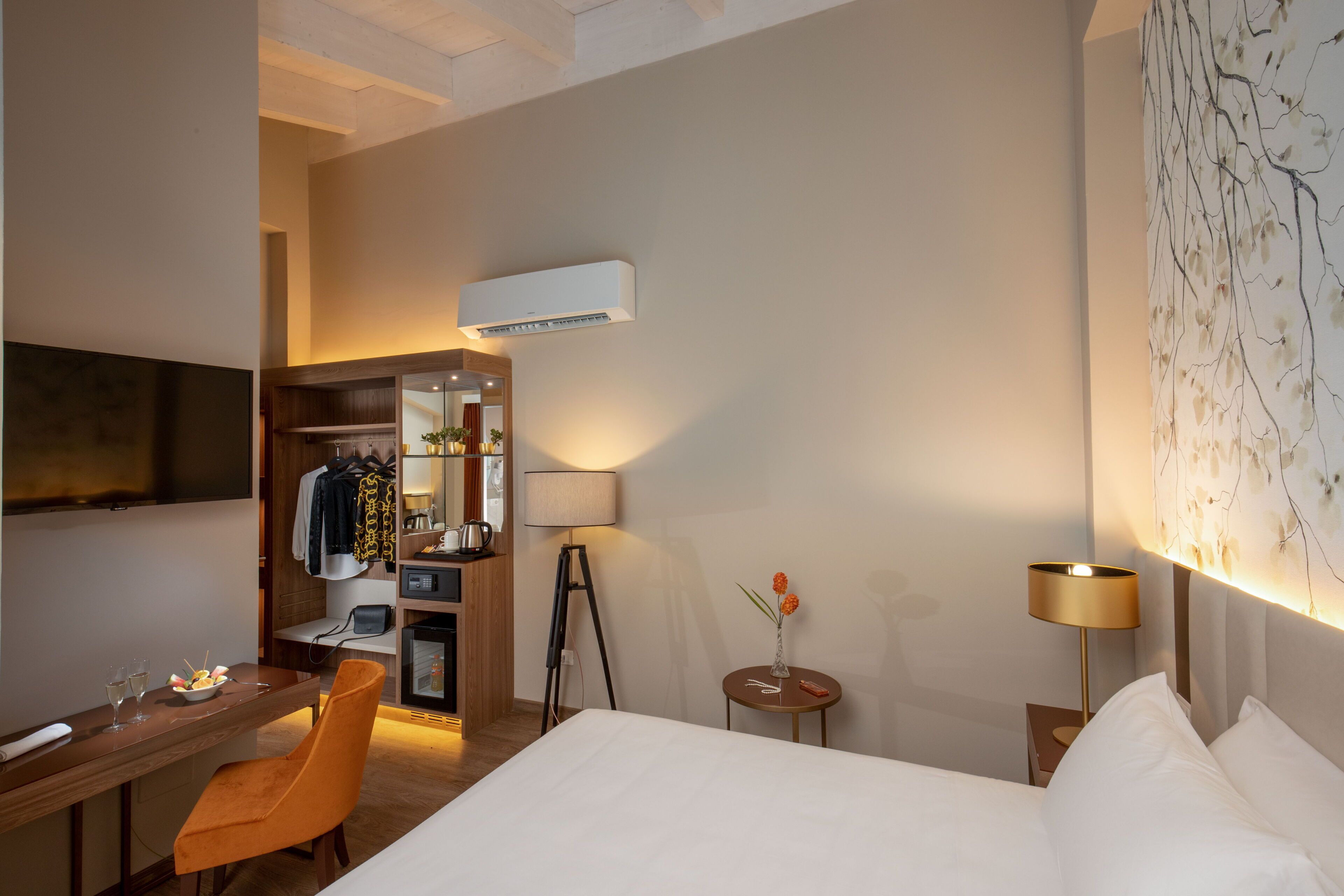 A hotel room with a bed, a desk, a chair, a small table with a vase, a lamp, and a wall-mounted air conditioner.
