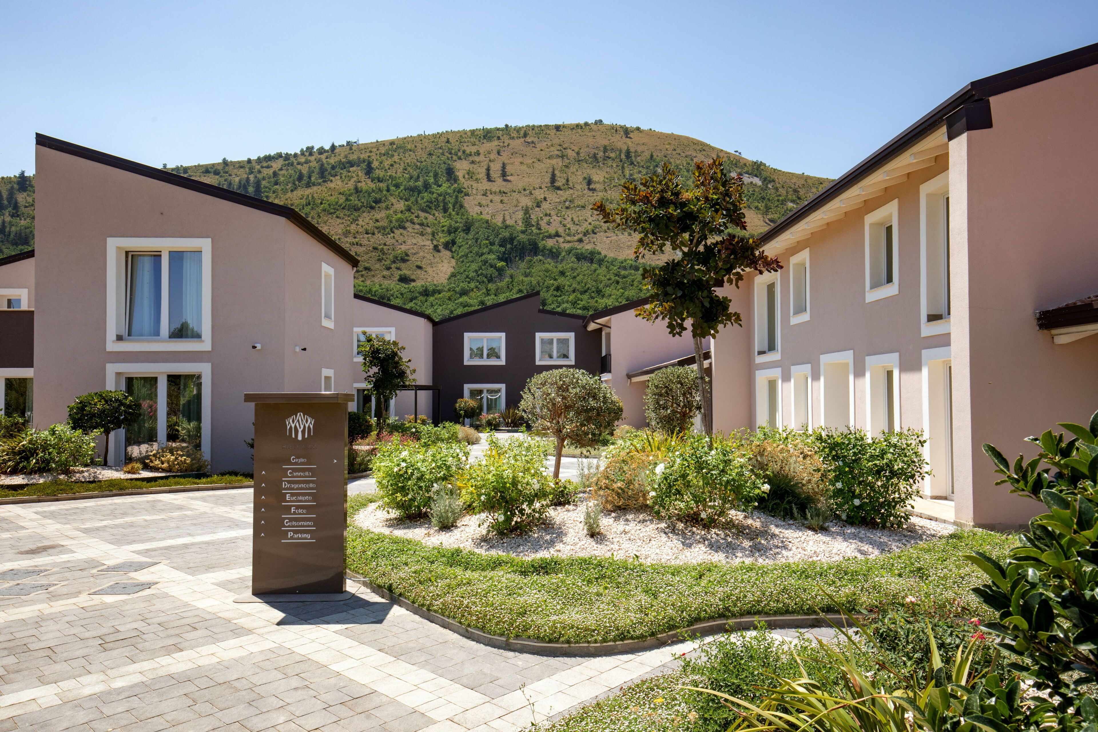 Un hotel moderno con un jardín ajardinado, un cartel que enumera los servicios y un telón de fondo montañoso.