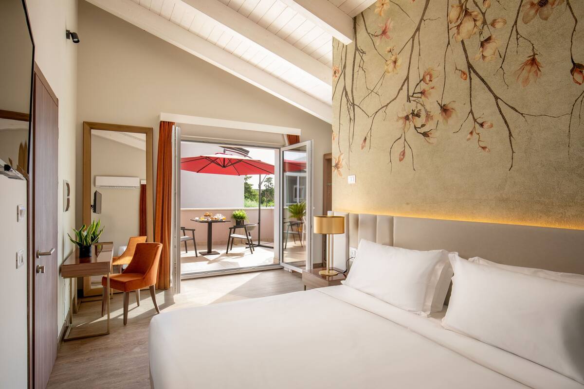 Quarto de hotel com uma cama grande, uma escrivaninha com cadeira, uma mesinha com cadeira e uma varanda com guarda-sol vermelho.