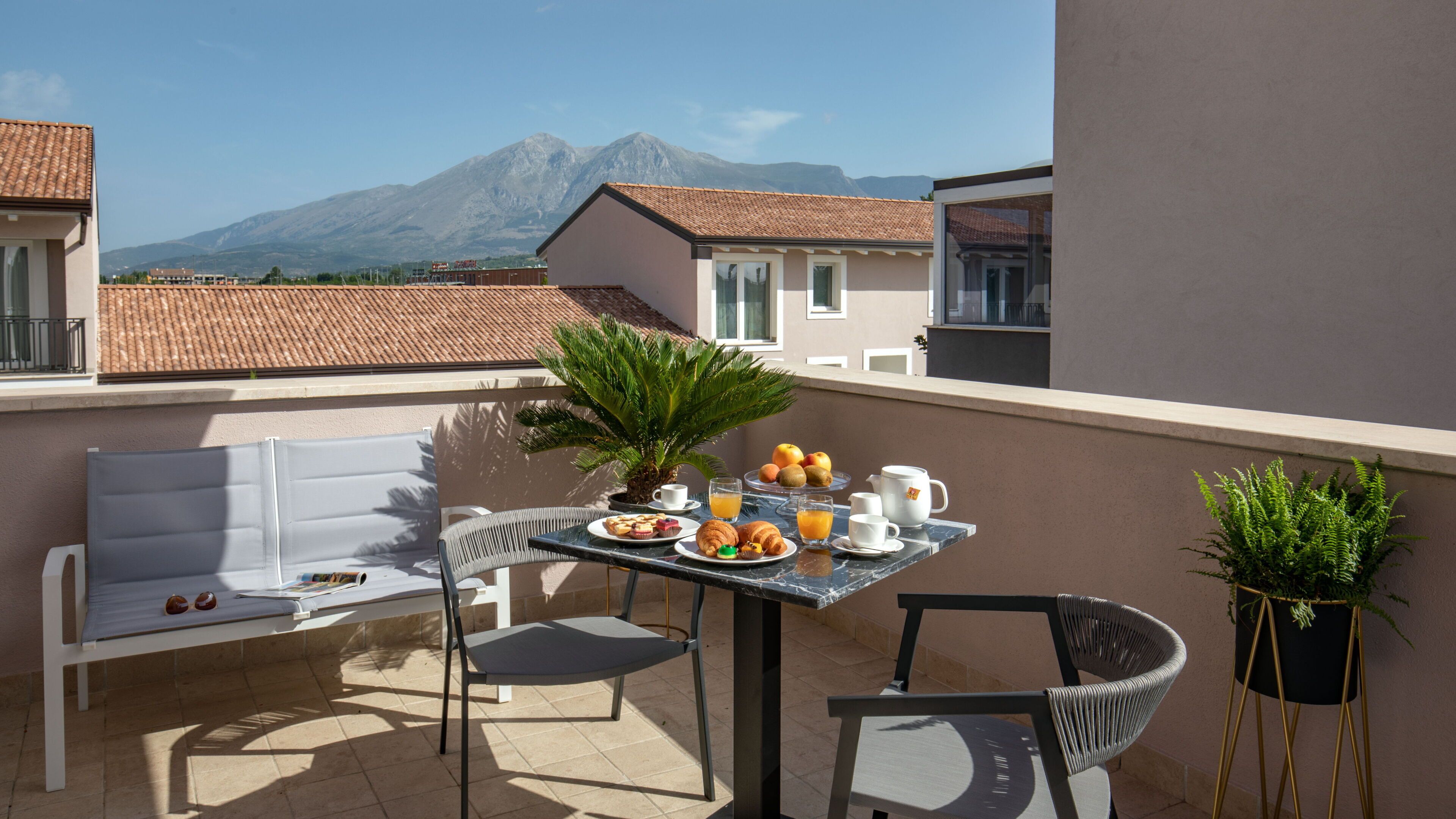 A balcony with a table set for breakfast, including croissants, fruit, and coffee. There are two chairs and a small potted plant.