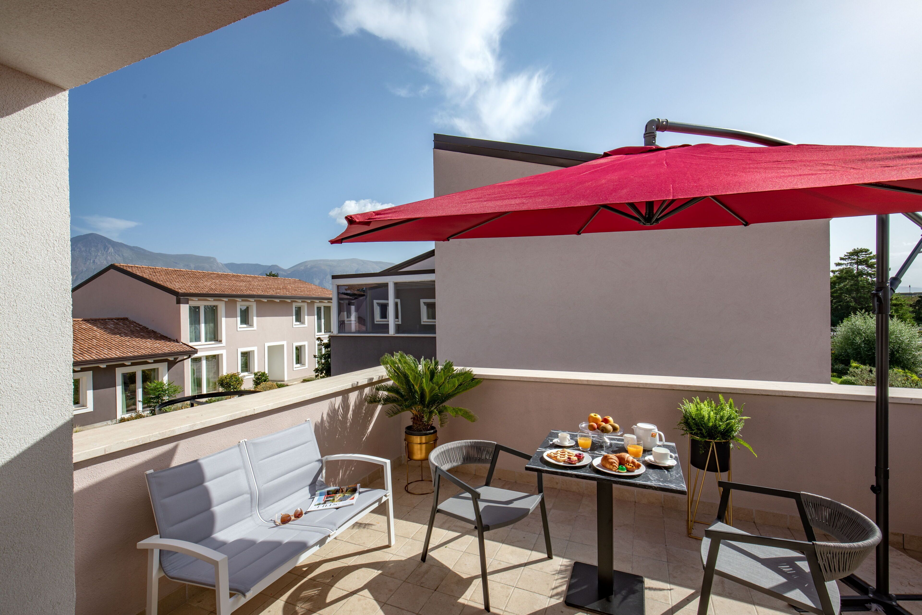 A balcony with a table set for breakfast, a red umbrella, and a white sofa.