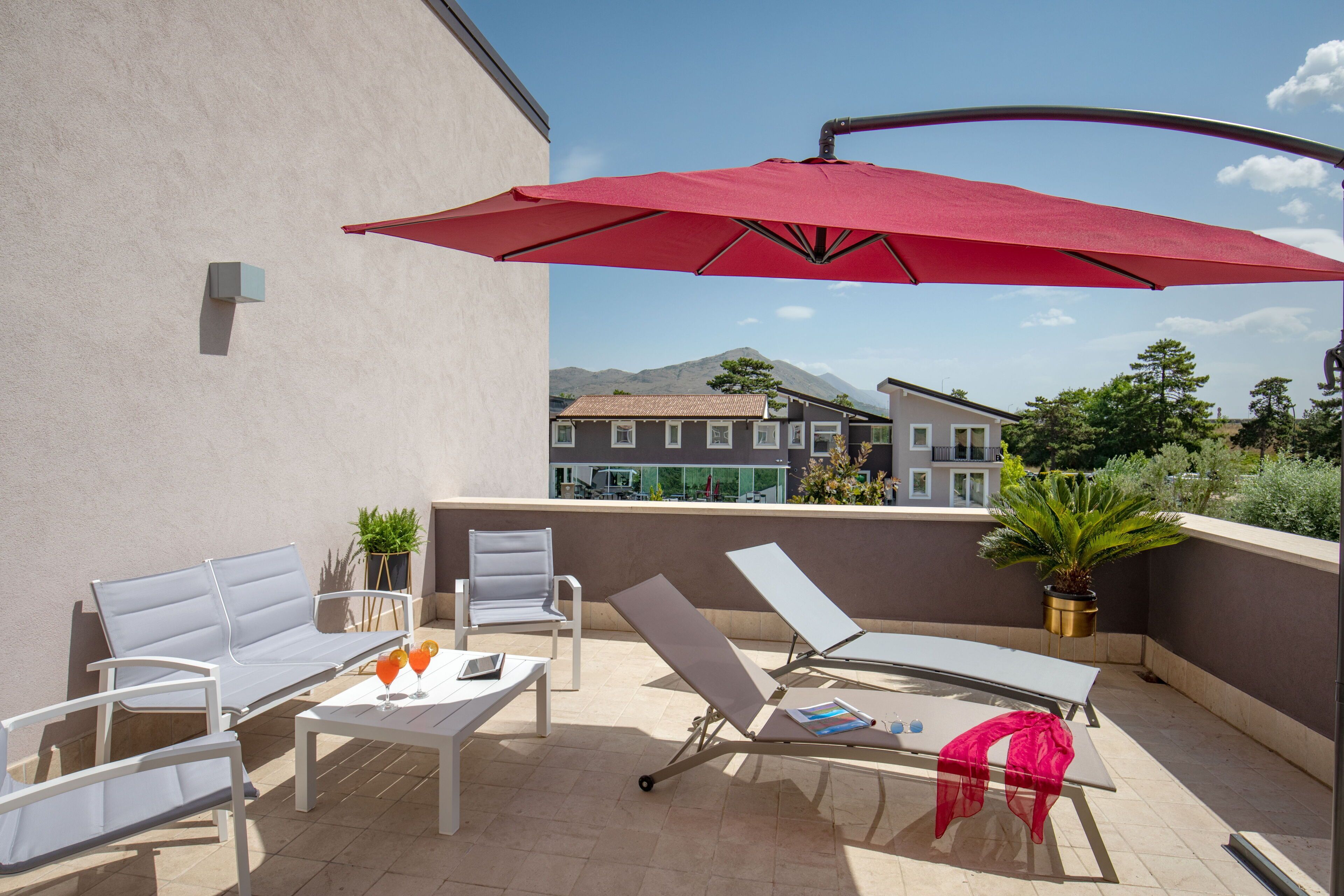Eine Dachterrasse mit weißen Liegestühlen, einem Tisch mit Getränken und einem roten Sonnenschirm.