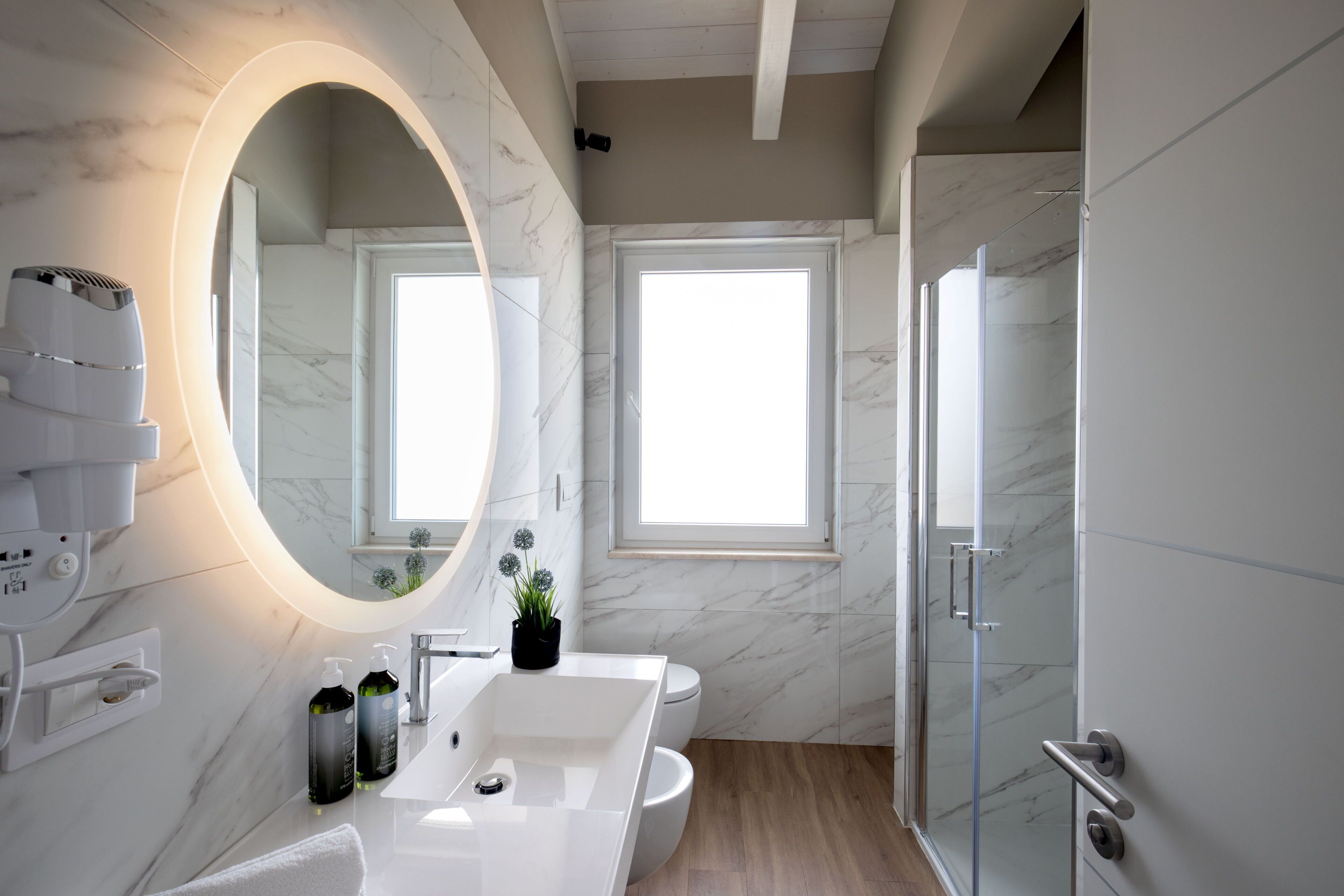 Un baño moderno con un espejo ovalado grande, un lavabo blanco, un inodoro y una cabina de ducha con mampara de vidrio.