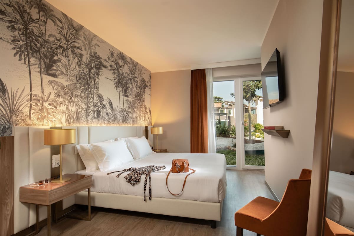 Quarto de hotel com uma cama grande, mesa de cabeceira com luminária, TV na parede e vista para o exterior.