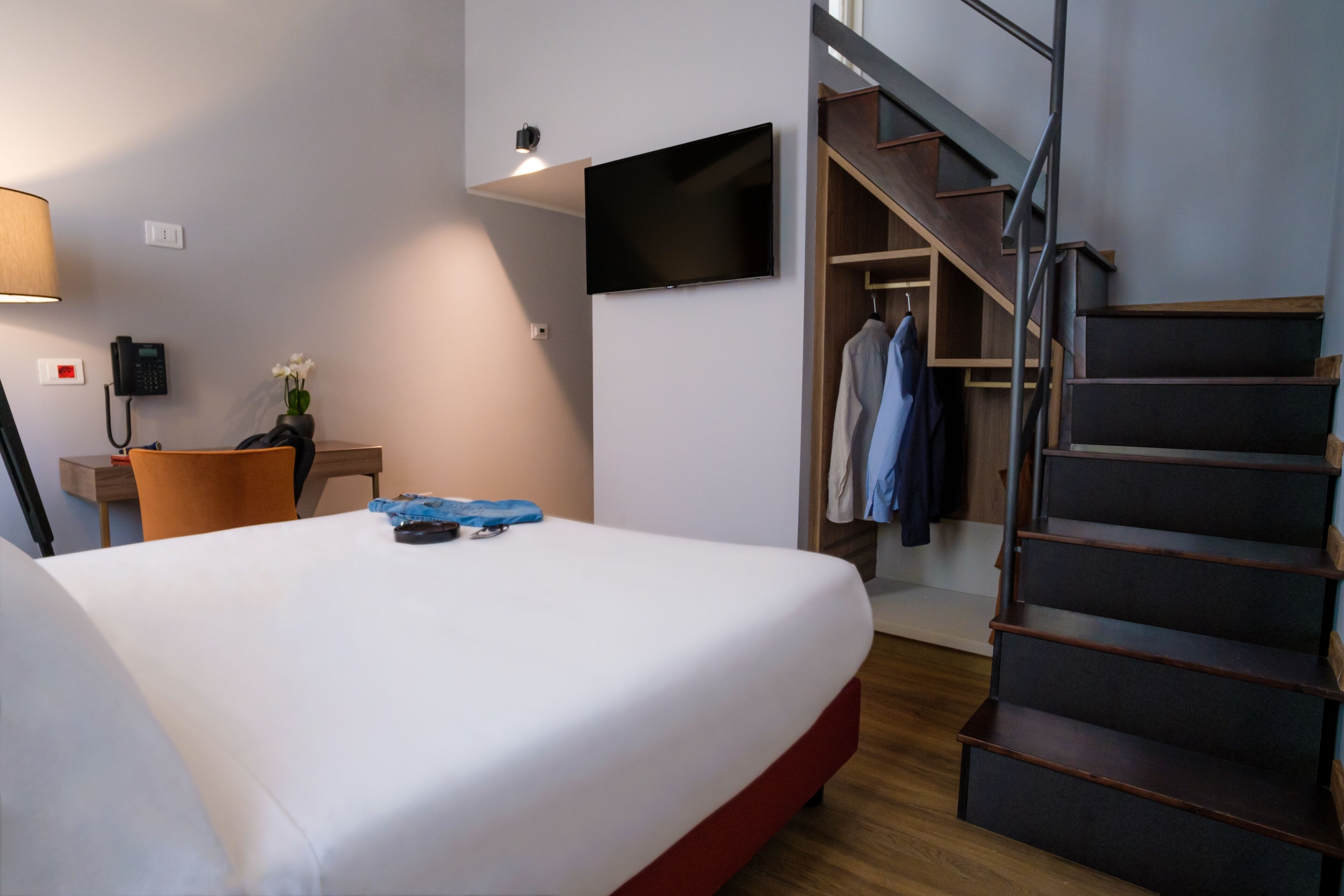 A hotel room with a bed, a desk with a lamp, a phone, a TV mounted on the wall, and a staircase.