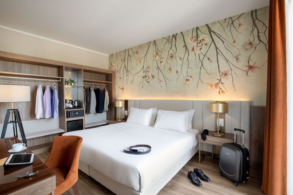 Quarto de hotel com uma cama grande, uma secretária com cadeira, um guarda-roupa e uma luminária na parede.